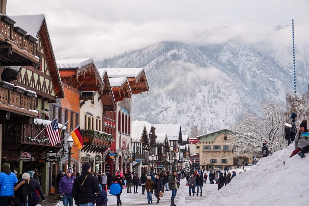 Snow-covered street in a Bavarian-style village with colorful buildings, flags, and people walking. Mountains in the background create a cozy winter scene.