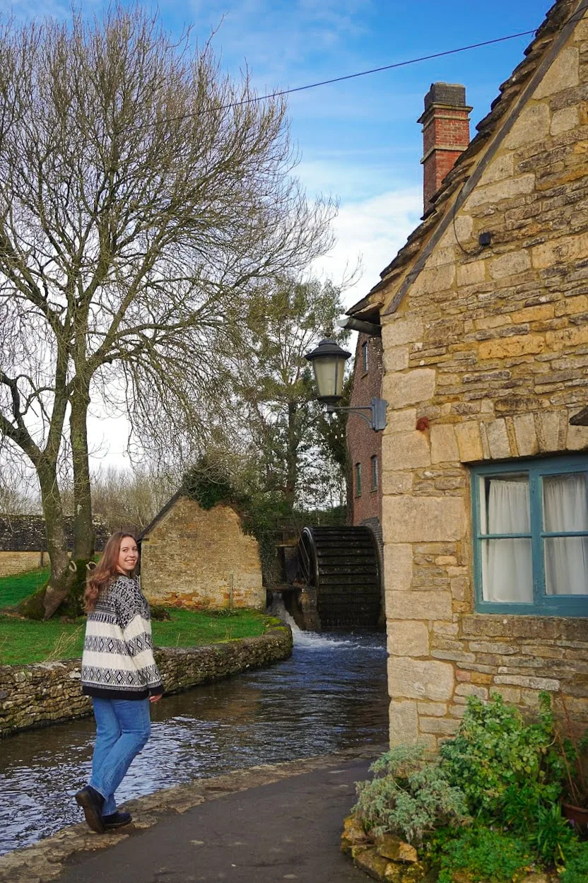 A woman in a patterned sweater and jeans stands by a small river near a rustic stone building with a blue window. The scene is peaceful and picturesque.