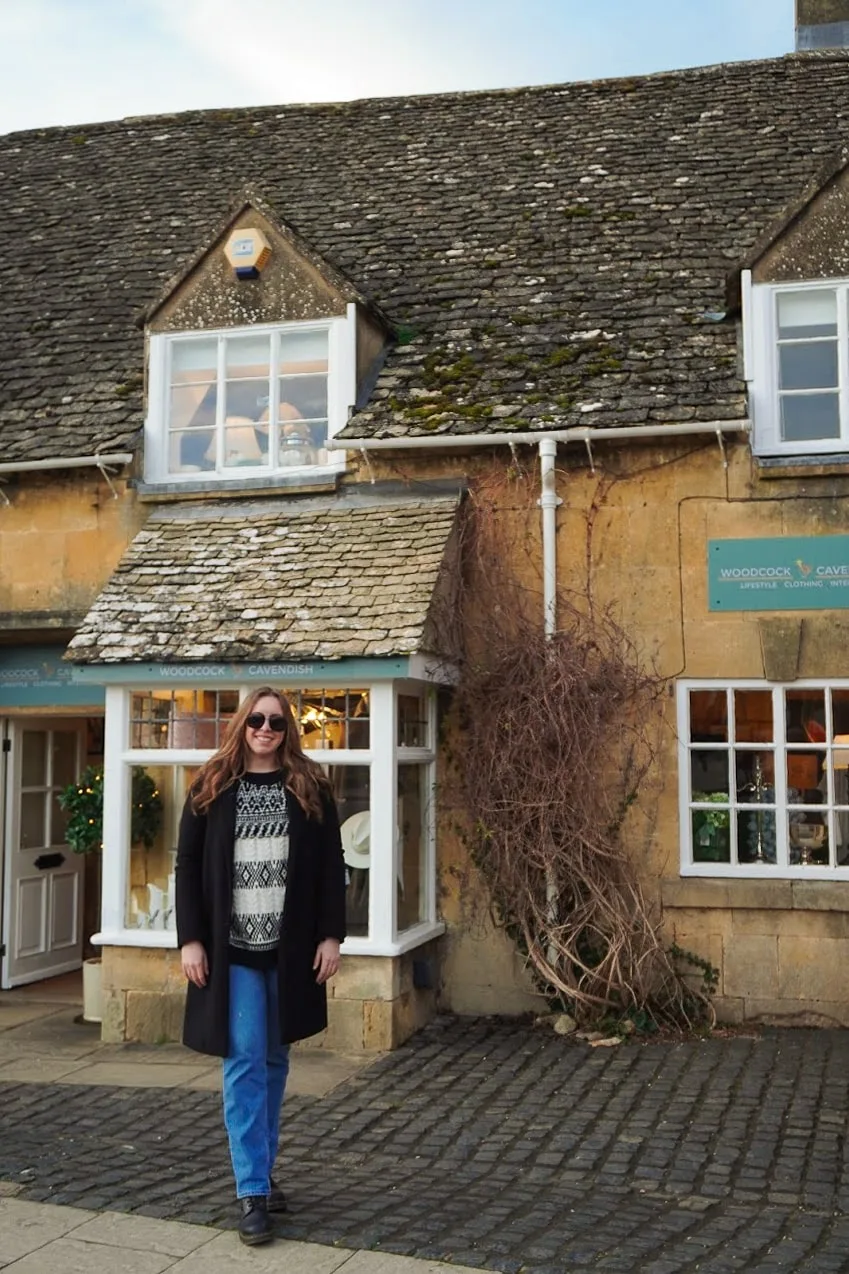 A woman in sunglasses stands smiling in front of a quaint stone cottage with a mossy roof and small shop windows. The atmosphere is cozy and rustic.