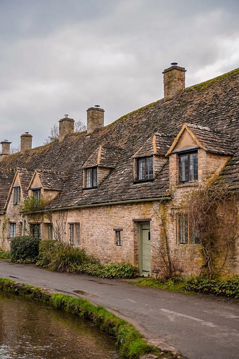 A row of charming stone cottages with steep, shingled roofs and small dormer windows line a quiet canal, evoking a serene, rustic atmosphere.