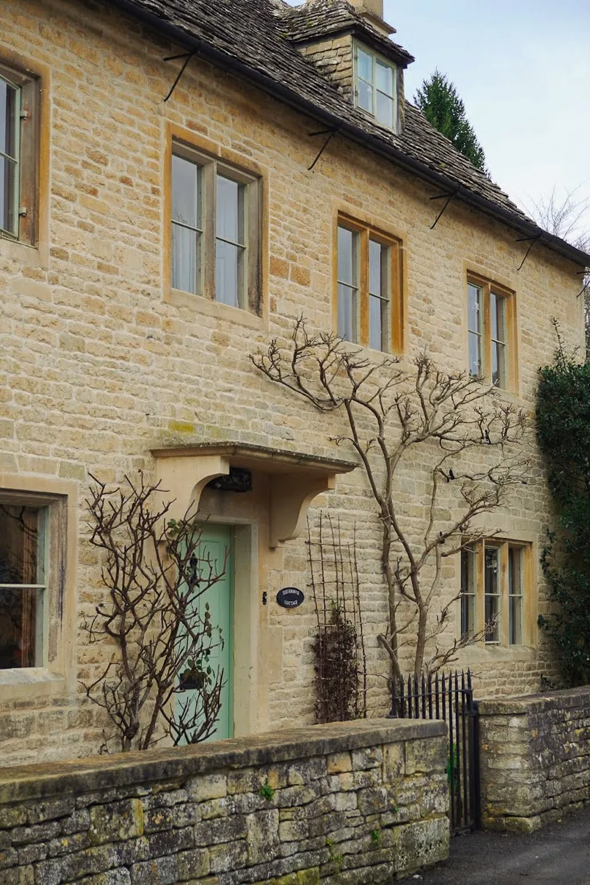 Stone cottage in Cotswolds AONB with pale green door, bare branches climbing the walls. Rustic charm and serene atmosphere, enclosed by a low stone wall.
