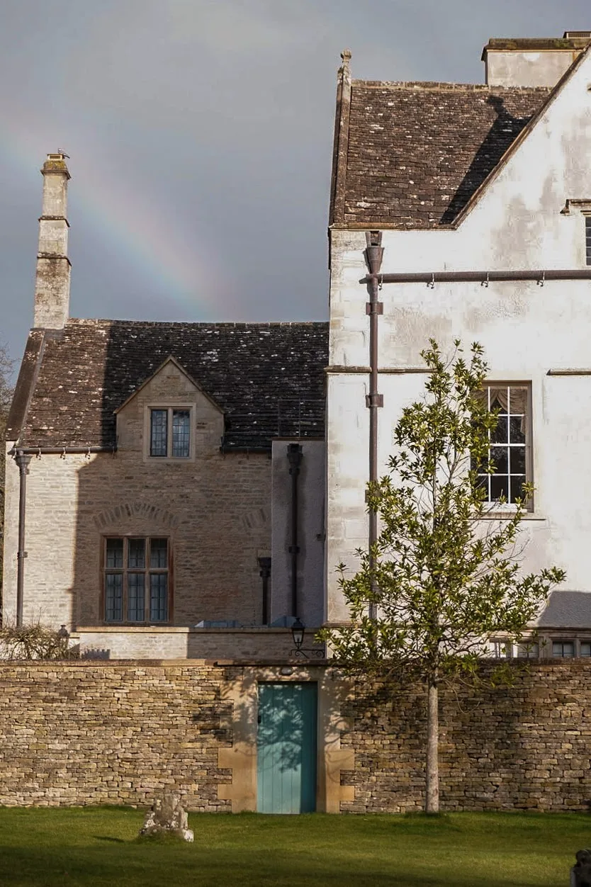 A rustic stone building with a weathered roof sits under a cloudy sky. A faint rainbow arcs in the background above a garden with a green door and tree.