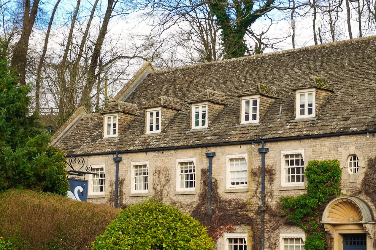 Cozy stone cottage with ivy-covered walls, dormer windows, and a rustic slate roof, surrounded by lush greenery. The scene feels quaint and serene.