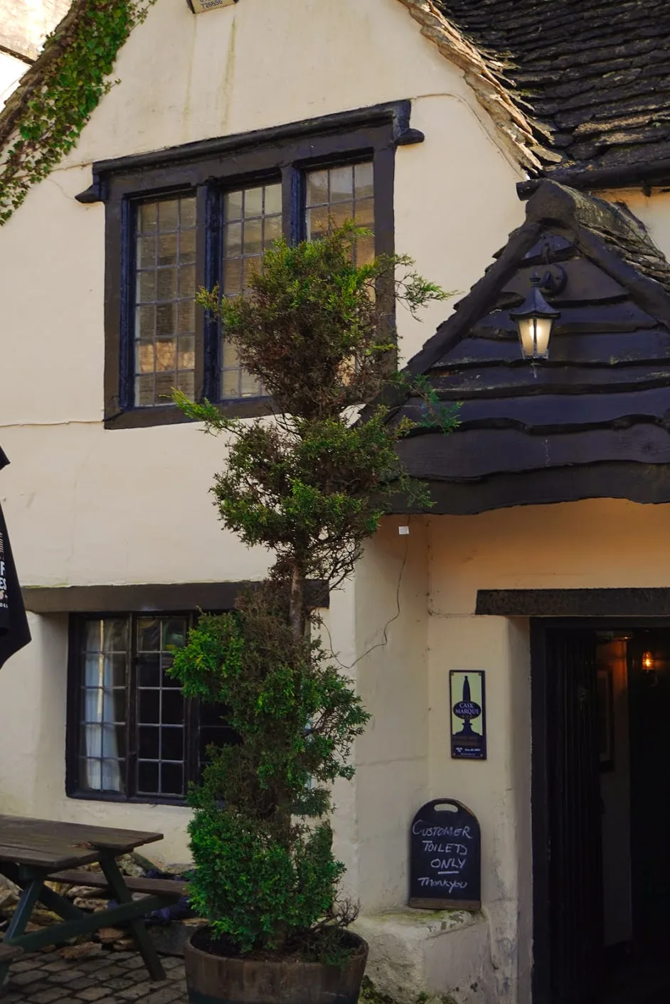 Old stone building with ivy, black wooden beams, and vintage lantern. A sign near the door reads "Customer Toilet Only." Cozy and quaint atmosphere.