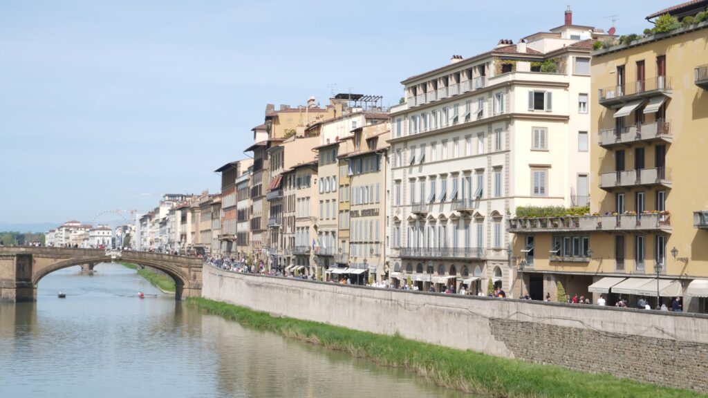 Scenic view of colorful historic buildings lining the Arno River in Florence, with a stone bridge and clear blue sky, creating a peaceful atmosphere.