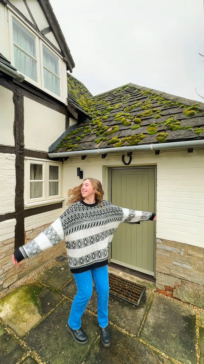 Young woman joyfully twirls on a paved patio in front of a rustic house. The roof is moss-covered, and she wears a cozy, patterned sweater.