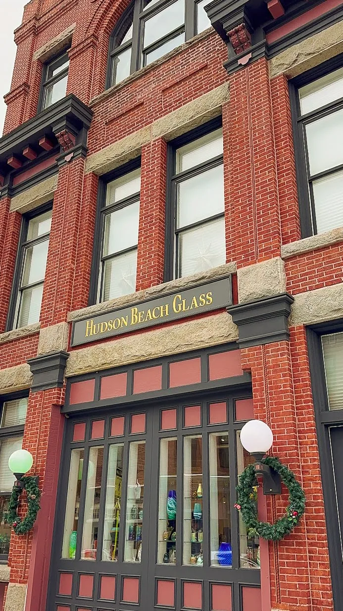 Historic red-brick building with Hudson Beach Glass sign. Large windows showcase colorful glass art. Festive wreaths hang on either side of the entrance.