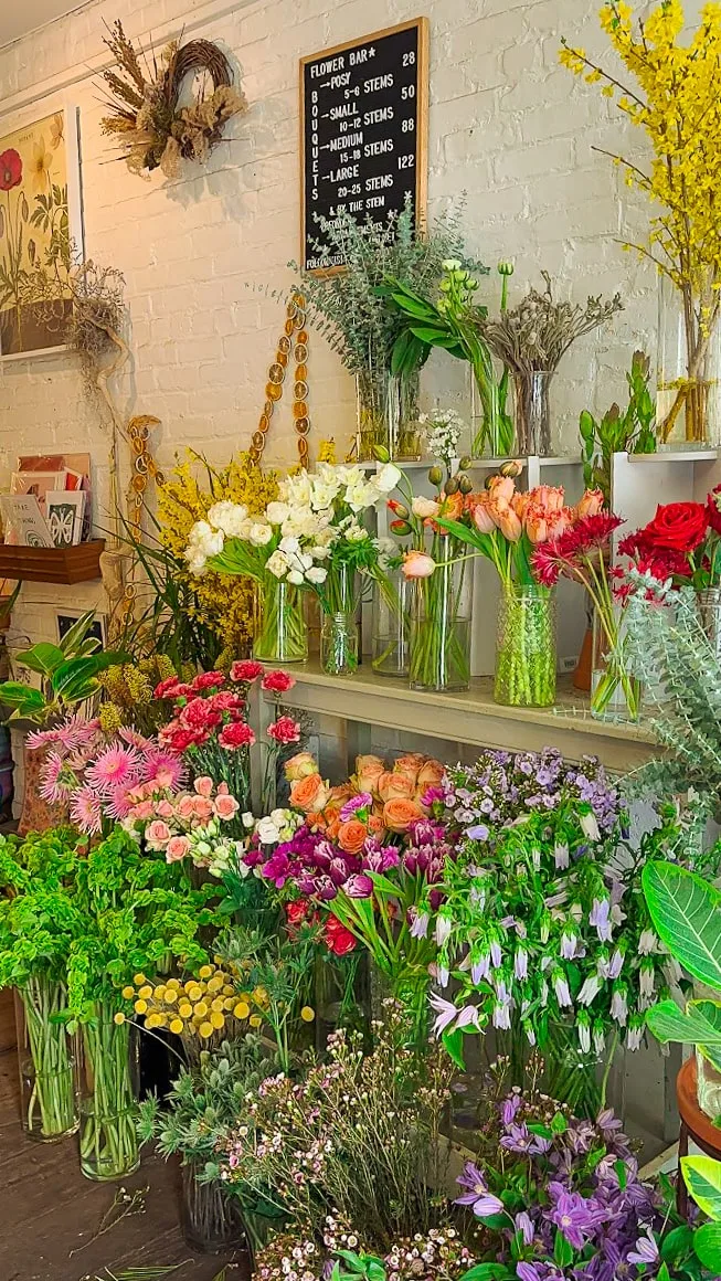 A vibrant flower shop displays assorted blooms, including roses, tulips, and daisies, in vases against a white brick wall. A price board hangs above.