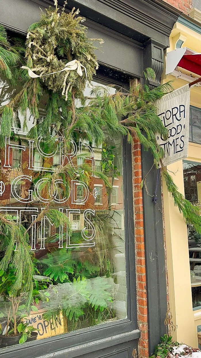 Shopfront with lush greenery: pine garland and wreath above a window displaying "Flora, Good Times." Inside, vibrant plants create a welcoming vibe.