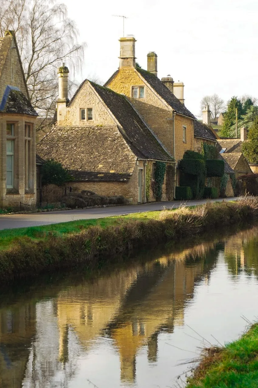 Rustic stone cottages with steep roofs line a quiet road beside a calm river. Reflections on the water create a serene, picturesque scene.