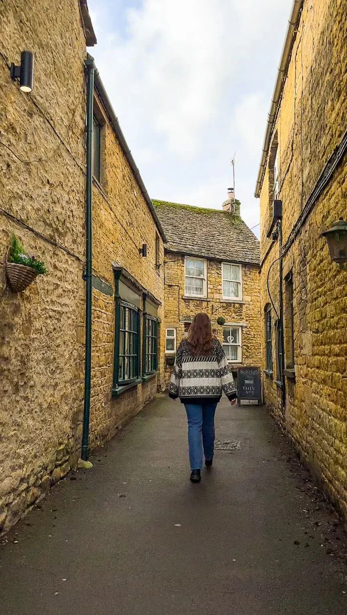 Person in a patterned sweater walks down a narrow, rustic alleyway lined with stone buildings. Atmosphere is quaint and peaceful in the Cotswolds AONB.