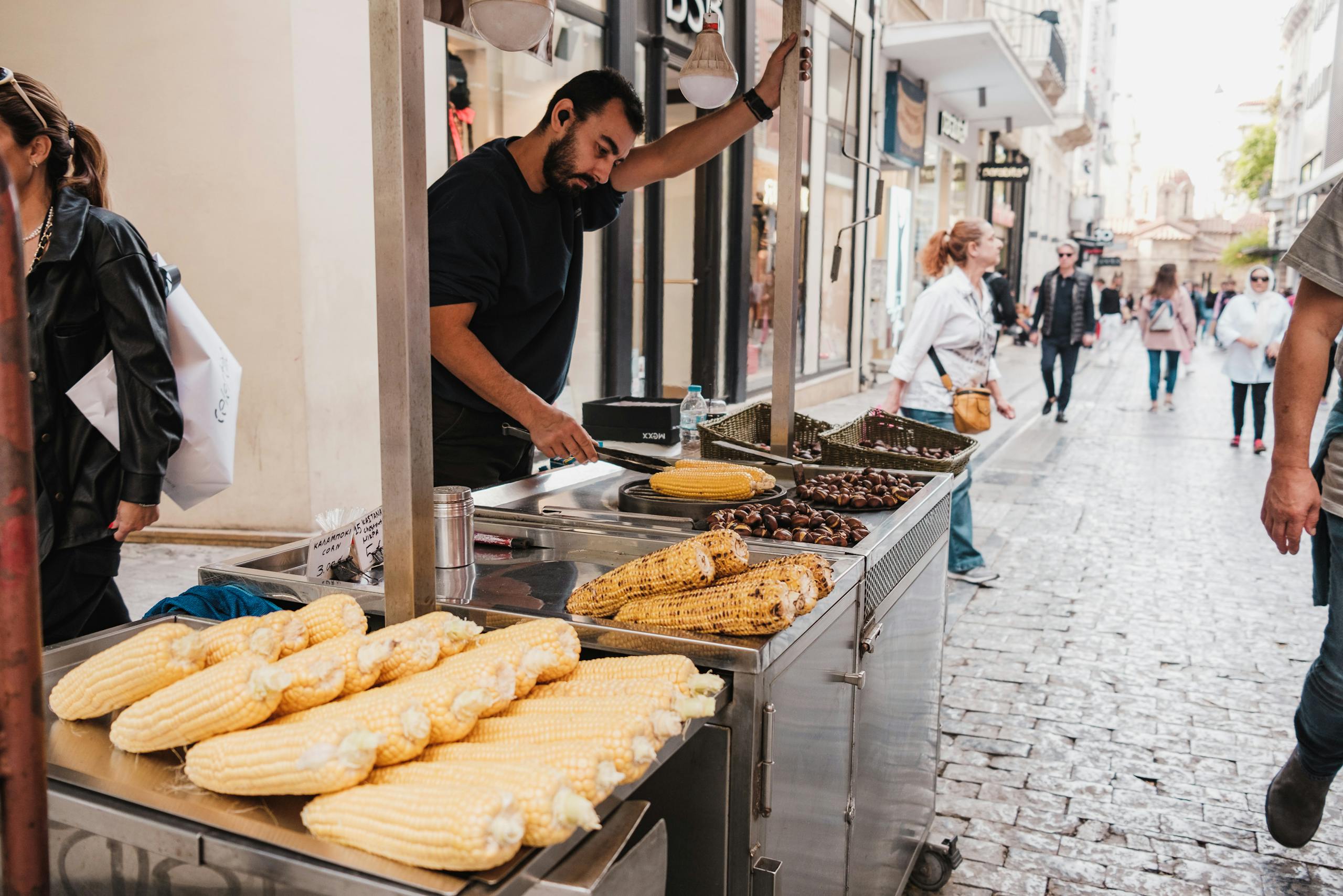 A lively street scene with a vendor selling grilled corn and chestnuts on a busy urban street.