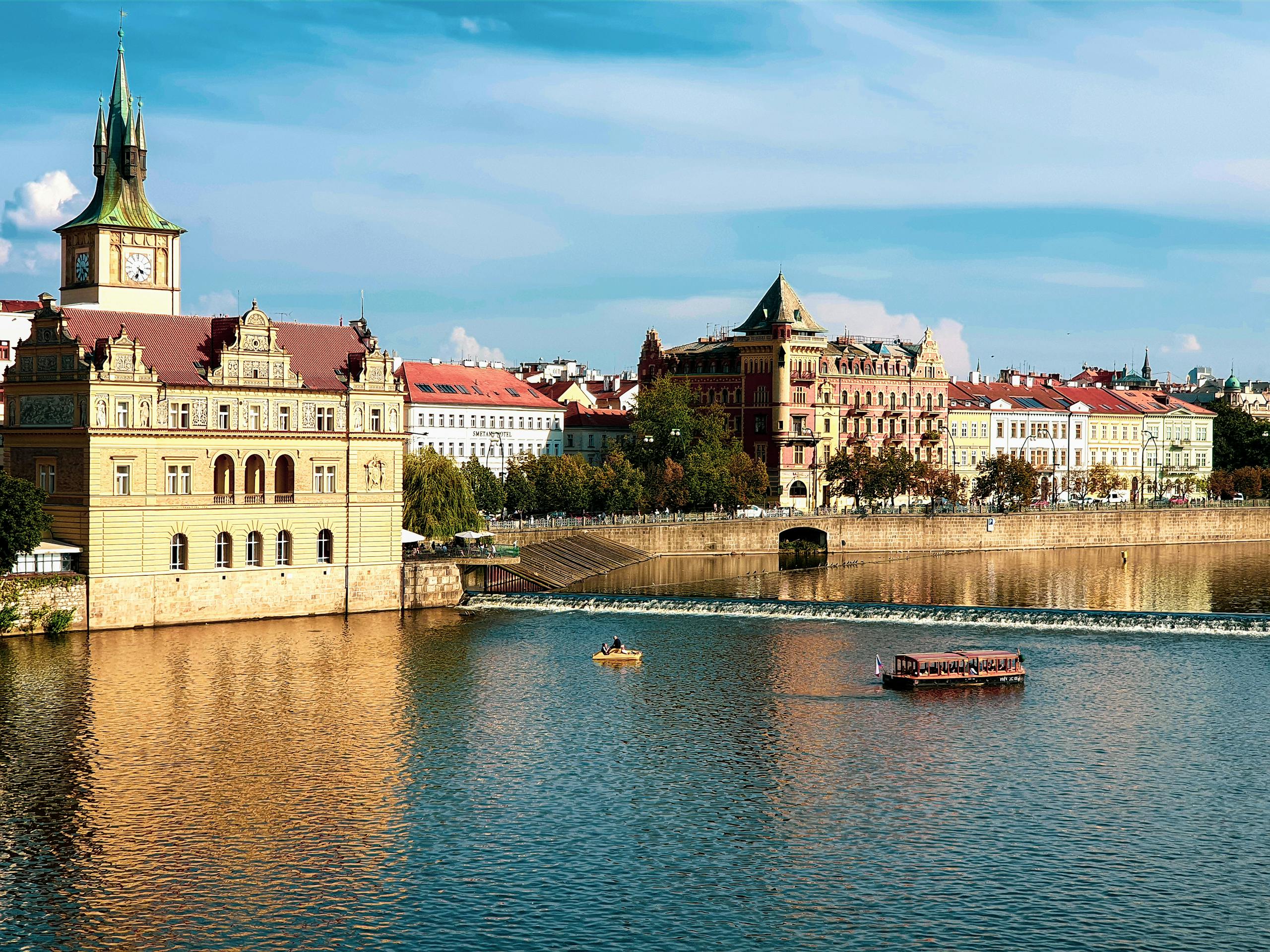 Historic buildings along a tranquil waterfront in Prague, Czechia, with clear skies.