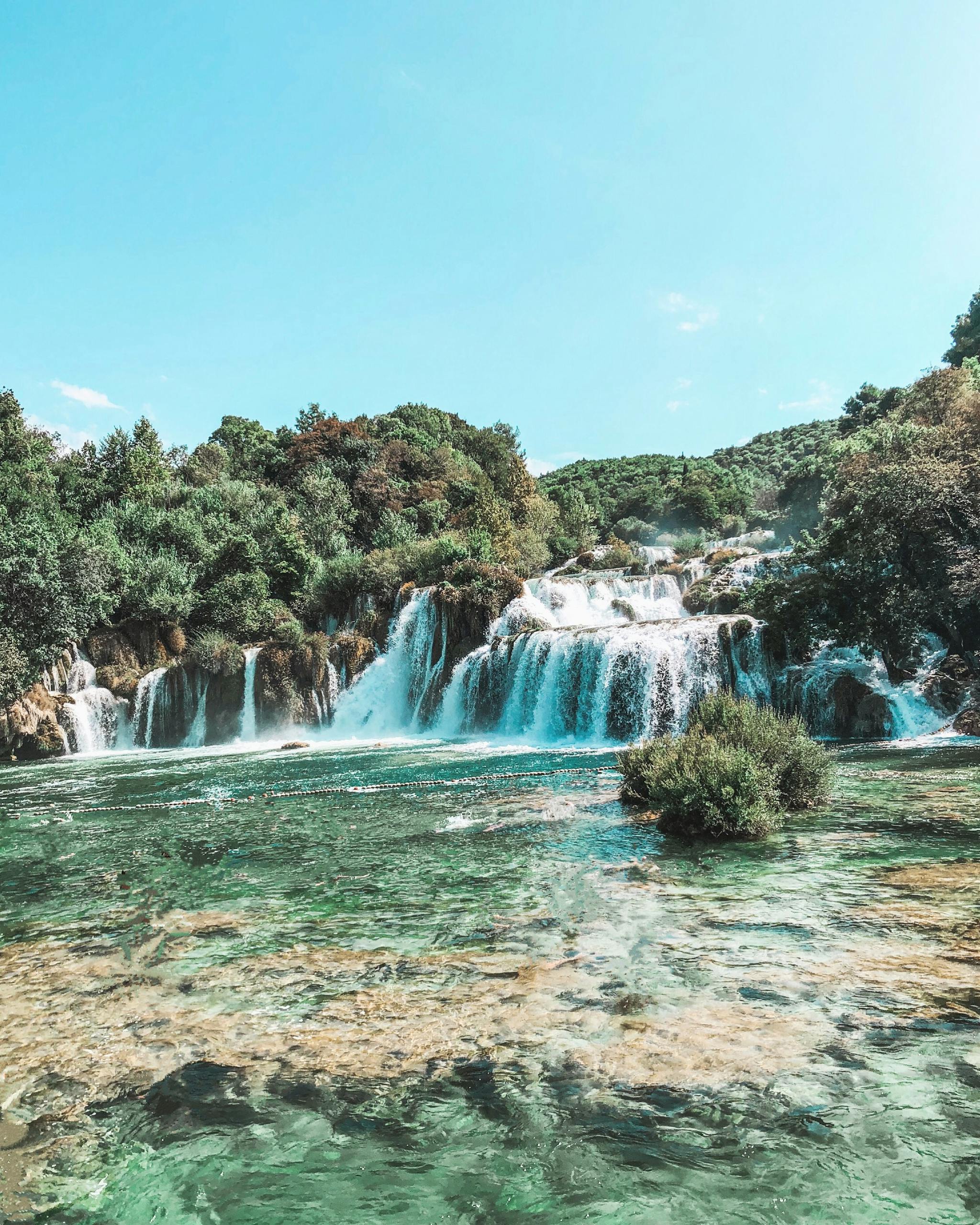 Scenic view of a waterfall cascading in Krka National Park, surrounded by lush greenery under a bright sky.