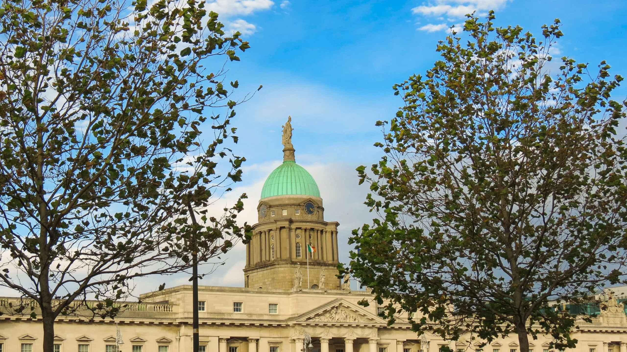 Dome of neoclassical building with a green roof, framed by two leafy trees. A statue tops the dome, beneath a bright blue sky dotted with clouds.