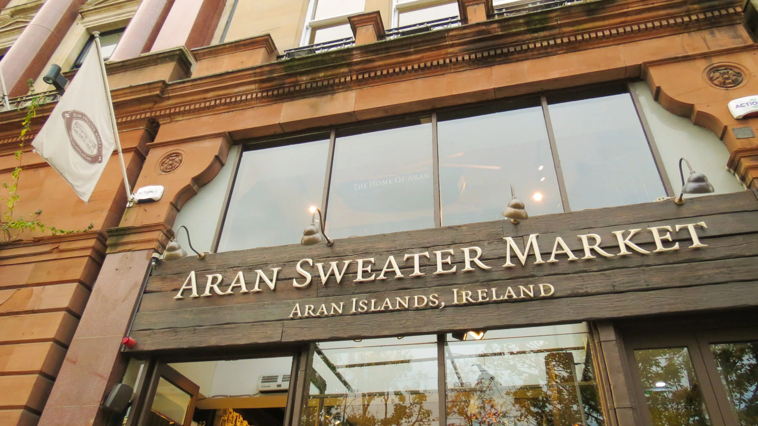 Front view of Aran Sweater Market with a flag on the left, nestled in the picturesque Aran Islands, Ireland. Beneath a wooden sign, the welcoming glass door invites you into this iconic spot, often highlighted in the best Ireland travel guides.