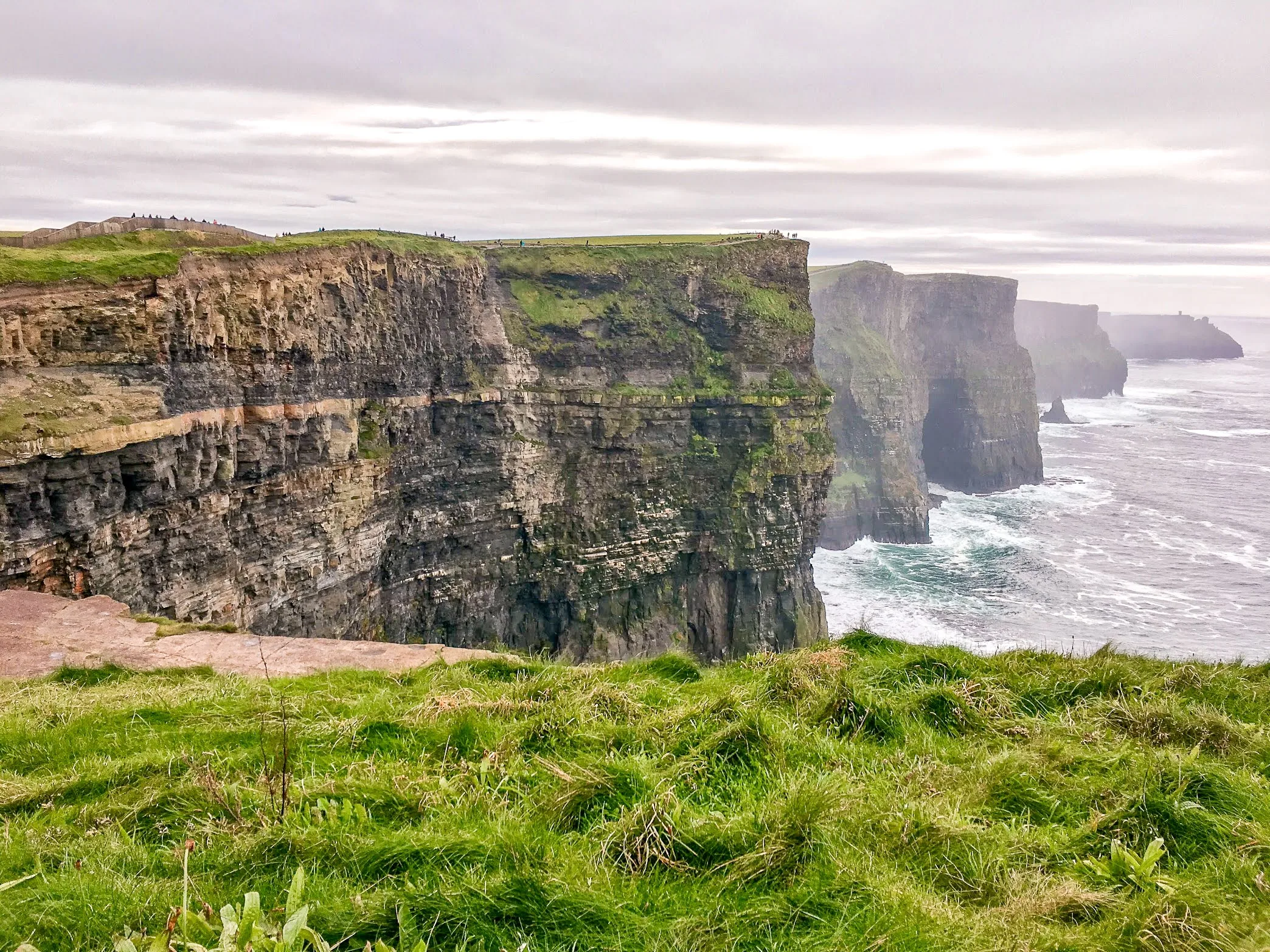 Cliffs of Moher on a cloudy day, featuring rugged, towering cliffs with layers of striated rock and green grass atop, meeting the crashing ocean below.
