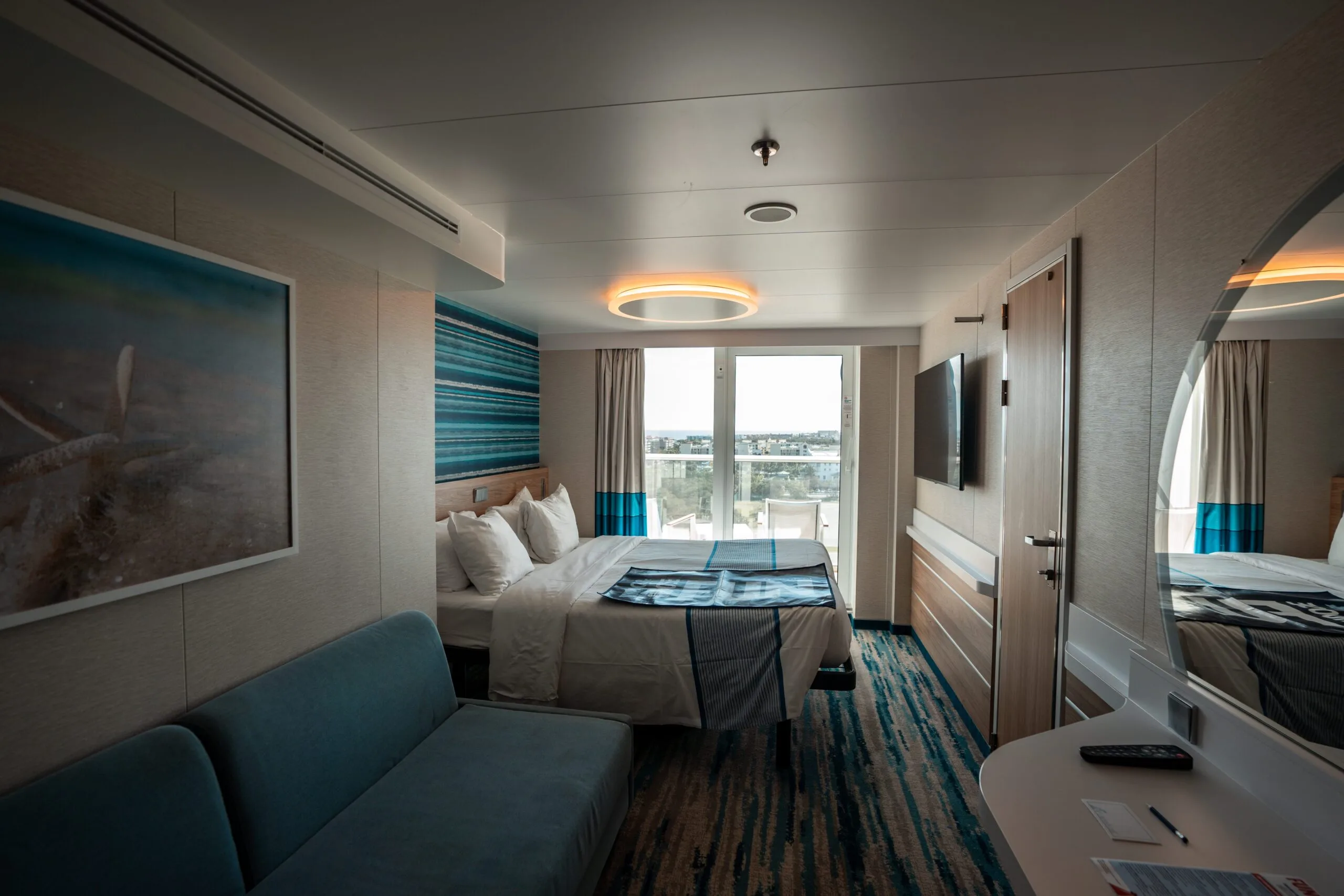 Modern hotel room with a queen bed, teal sofa, and abstract ocean artwork. A large window offers a city view. Warm lighting creates a cozy atmosphere.