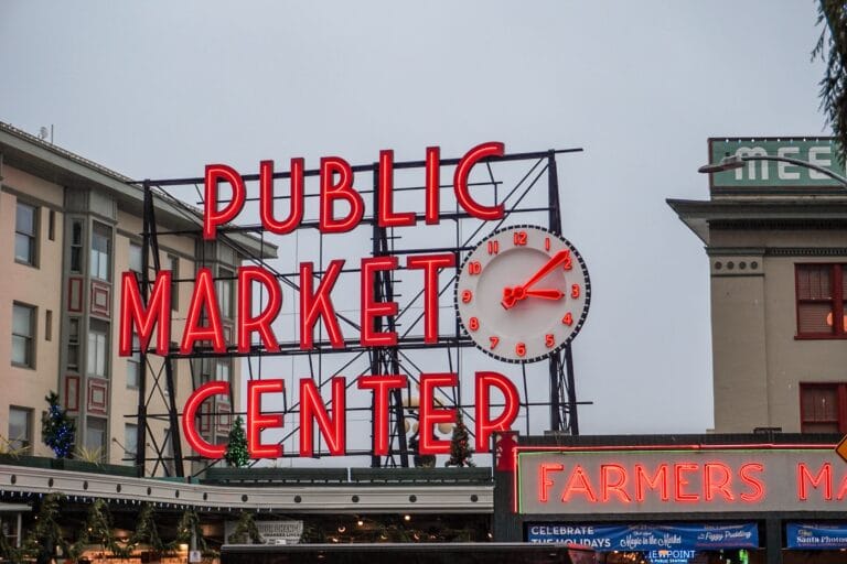 Bright neon sign reading "Public Market Center" with a red clock above. Below, another sign reads "Farmers Market." Urban and lively atmosphere.