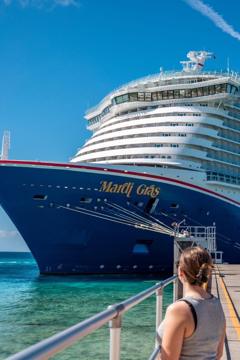 A person in a sleeveless top stands on a pier, gazing at a massive cruise ship named "Mardi Gras" against a clear blue sky. The scene conveys a sense of awe and anticipation.
