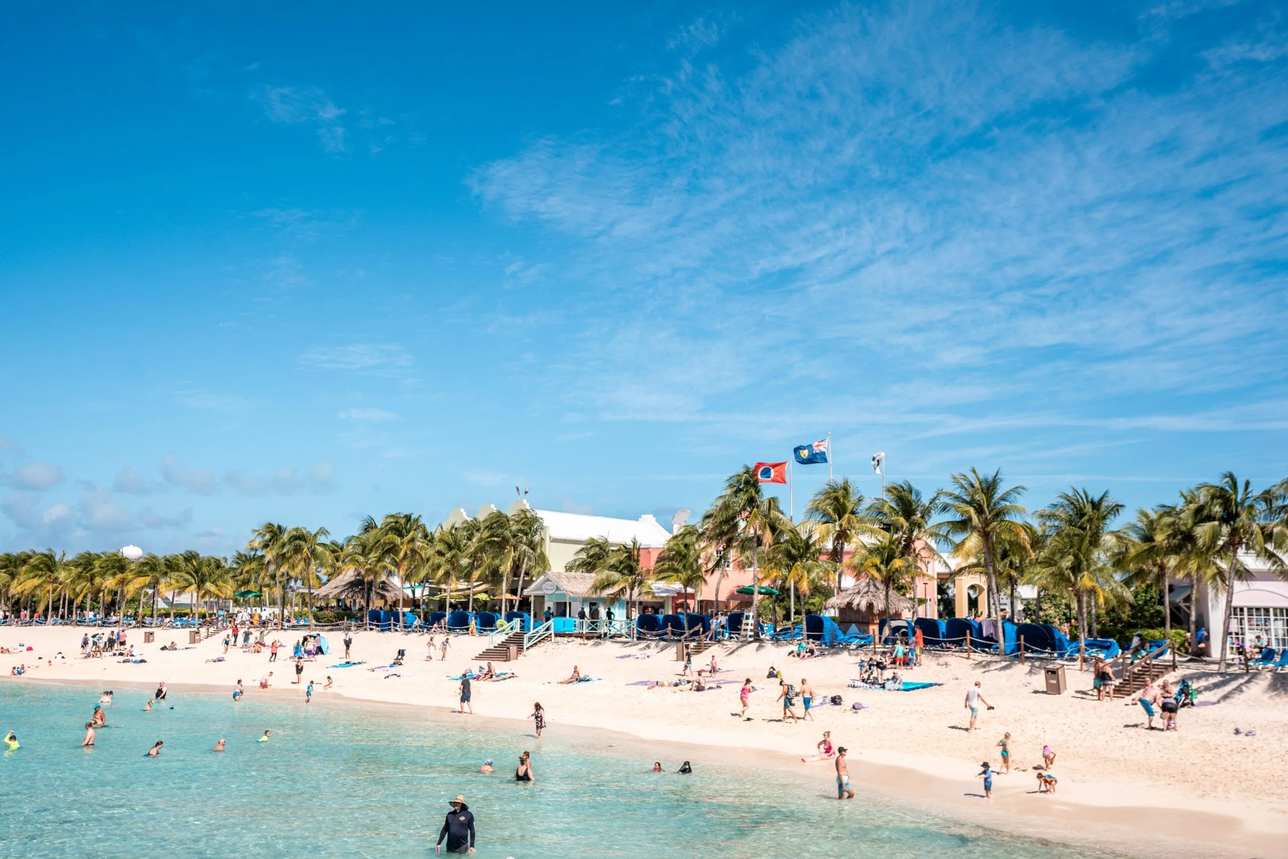 A sunny beach scene with turquoise waters, people swimming and relaxing on a sandy shore. Palm trees and colorful buildings line the background.