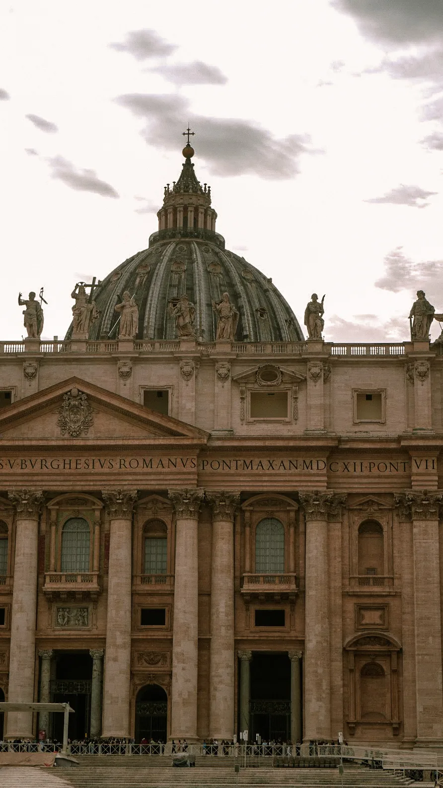 St. Peter's Basilica facade with grand columns and statues atop, under a cloudy sky. The dome is visible, conveying historical grandeur and solemnity.