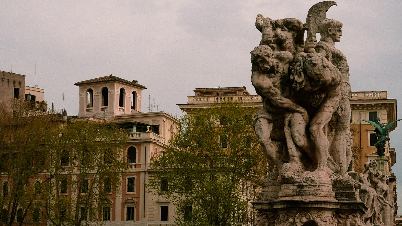 Stone statues of intertwined figures stand prominently against a backdrop of elegant, weathered buildings. A cloudy sky adds a timeless, historic feel.