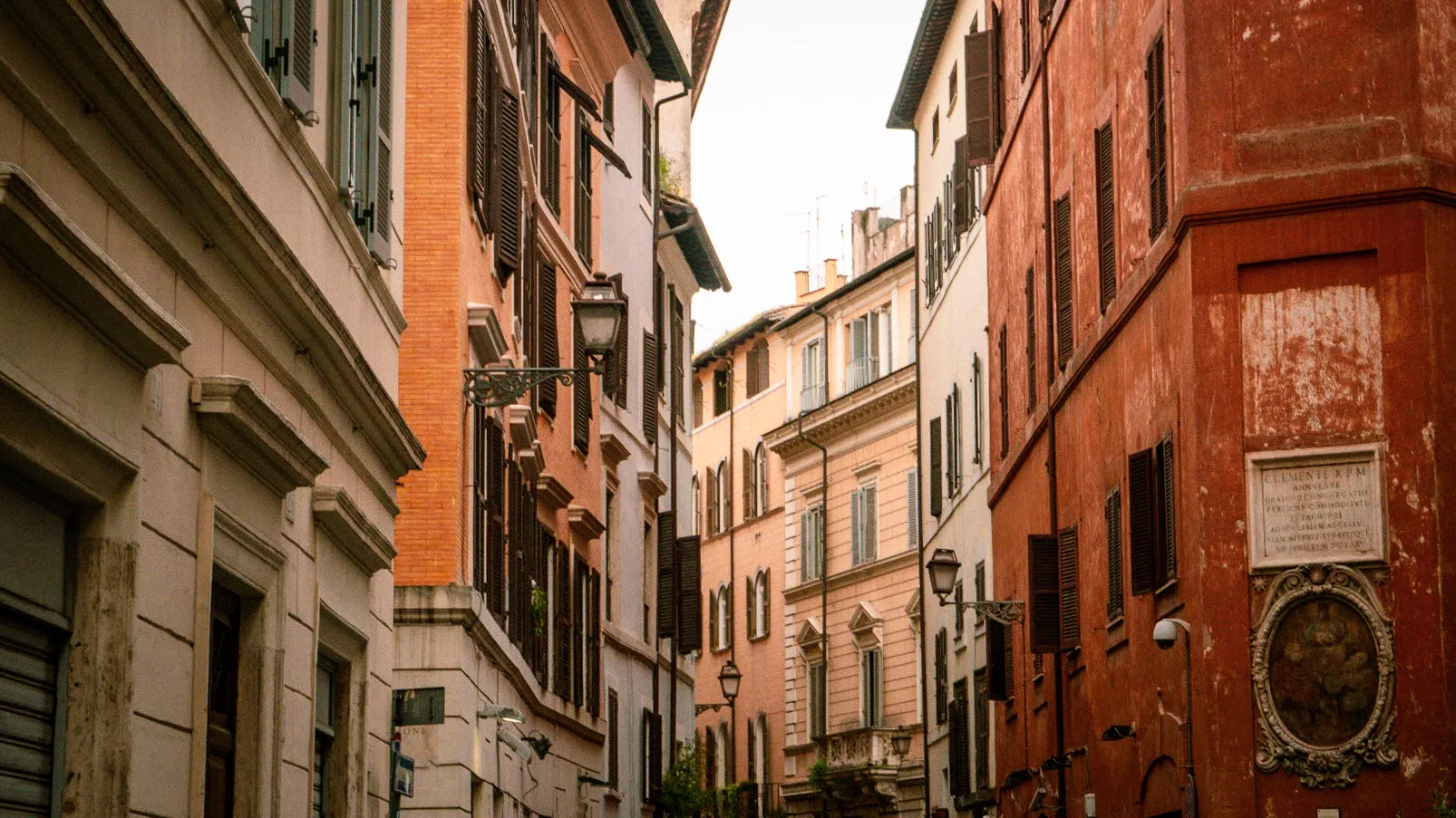 Narrow street lined with tall, old buildings featuring rustic red and orange facades, shuttered windows, and vintage street lamps, evoking a nostalgic ambiance.