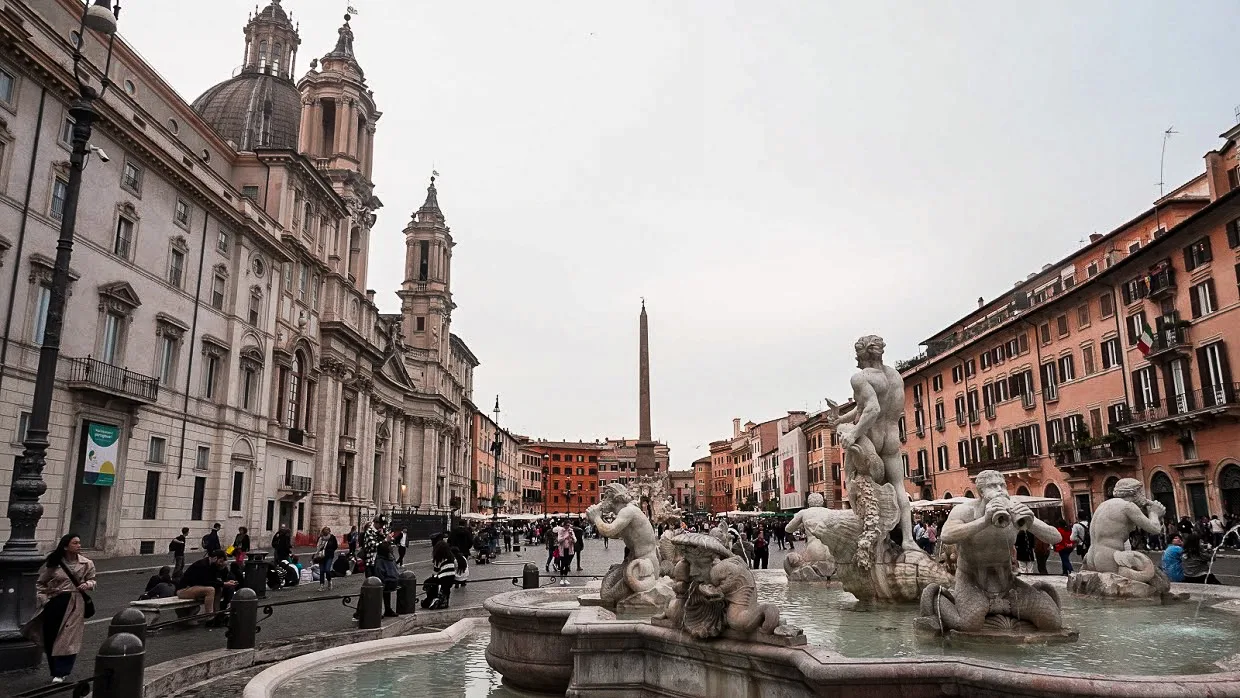 A bustling Piazza Navona in Rome, featuring a Baroque fountain with detailed sculptures. Historic buildings line the square, with an obelisk in the distance.