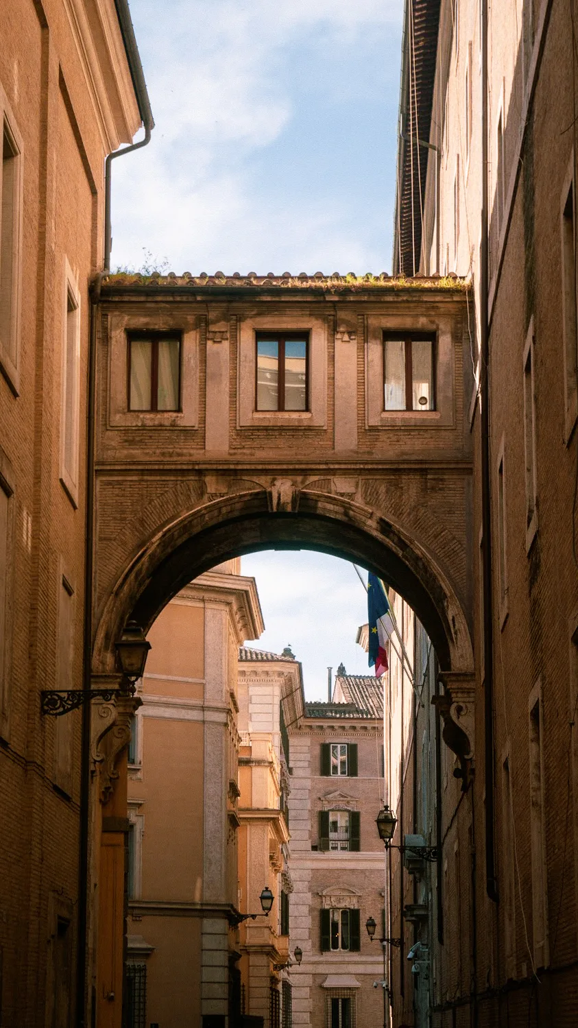 Narrow alley with an arched stone bridge connecting brick buildings, blue sky above. Italian flags visible, creating a historical, European ambiance.