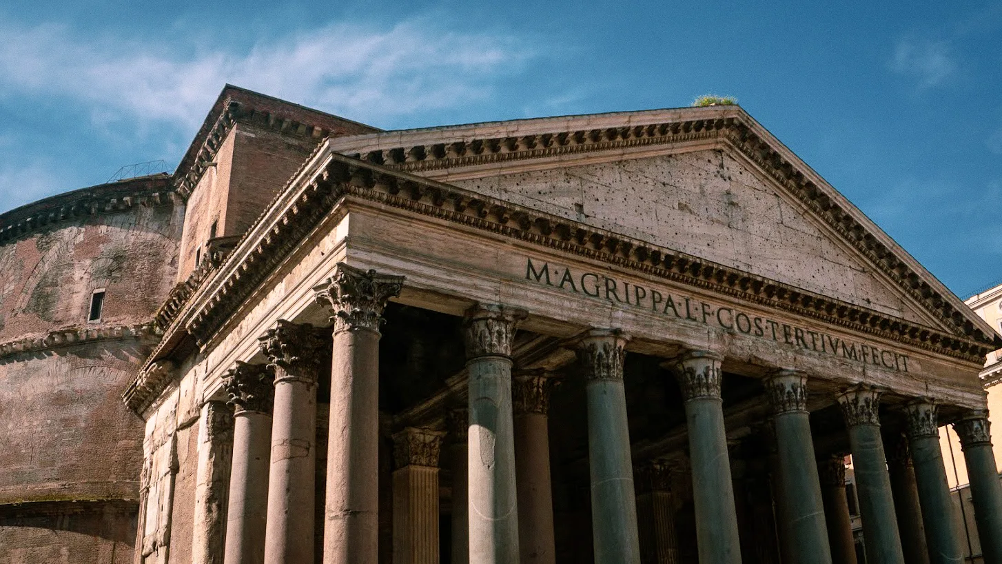 Ancient Roman building with weathered stone columns and triangular pediment under a clear blue sky. Latin inscription on the facade.