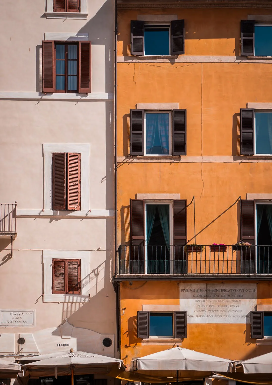 Colorful Italian buildings with wooden shutters and two-tone facades, one beige and one orange. A sunny ambiance with a bright sky enhances the vibrant scene.
