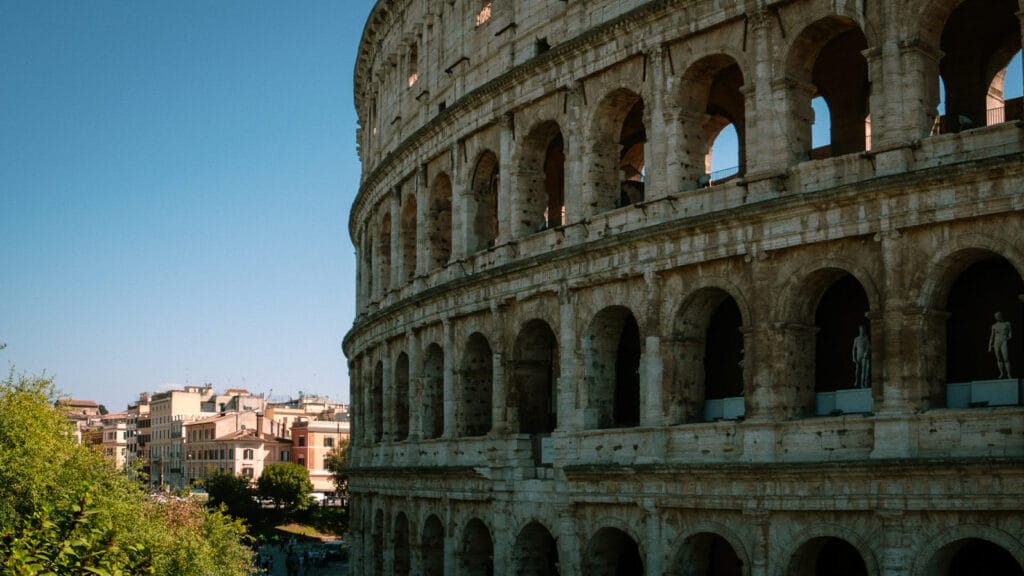 Close-up of the Colosseum in Rome, Italy, showing its ancient arches and statues under a clear blue sky. Nearby buildings add urban contrast.
