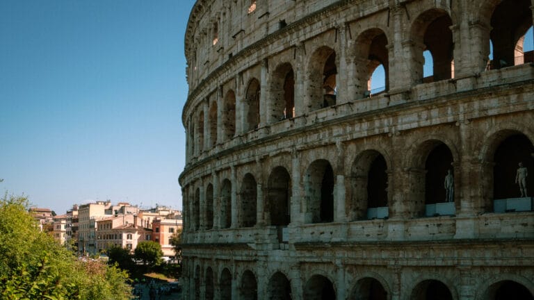 Close-up of the Colosseum in Rome, Italy, showing its ancient arches and statues under a clear blue sky. Nearby buildings add urban contrast.