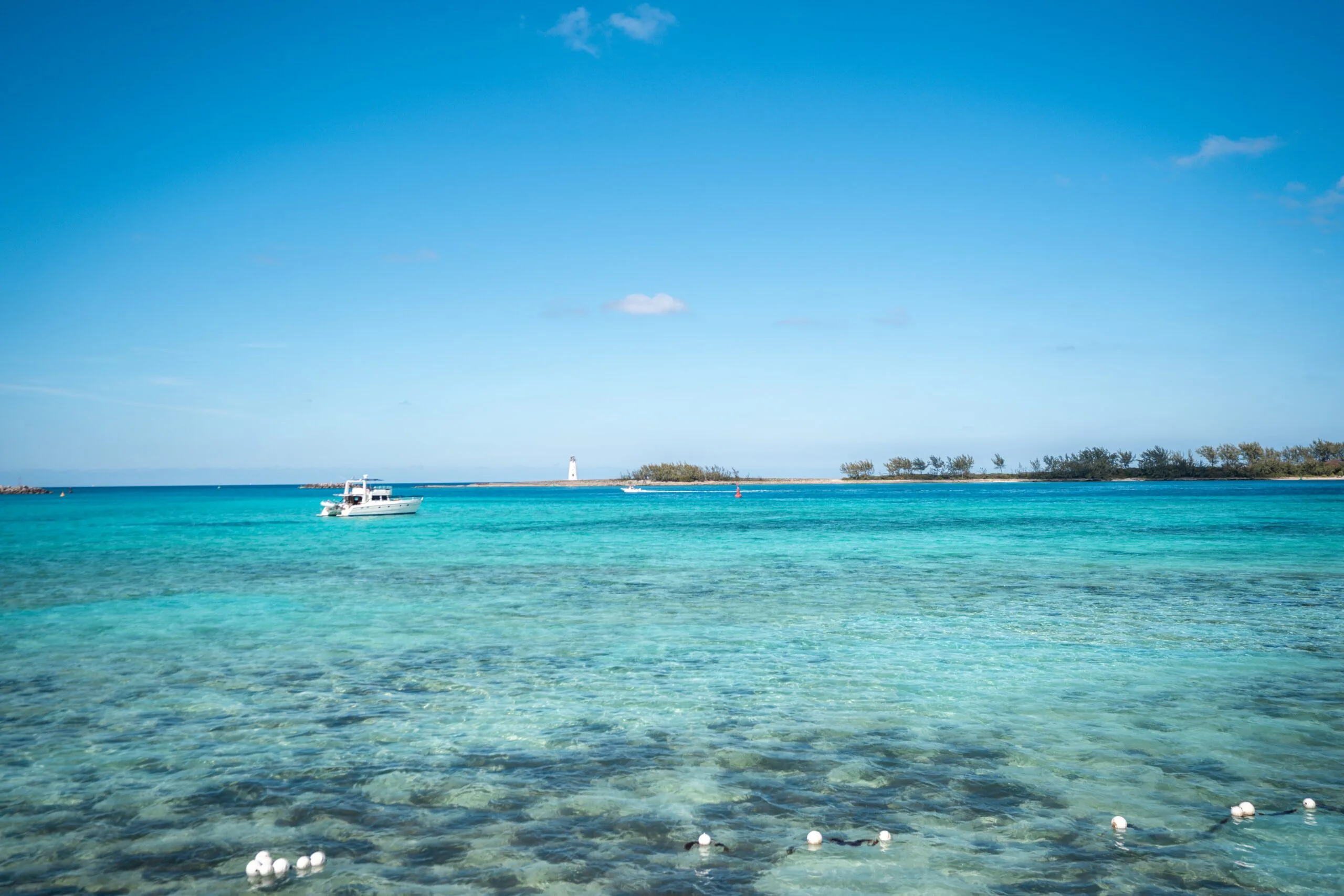 A boat sails on clear turquoise waters under a bright blue sky. A distant island with a lighthouse creates a serene and tranquil seascape.