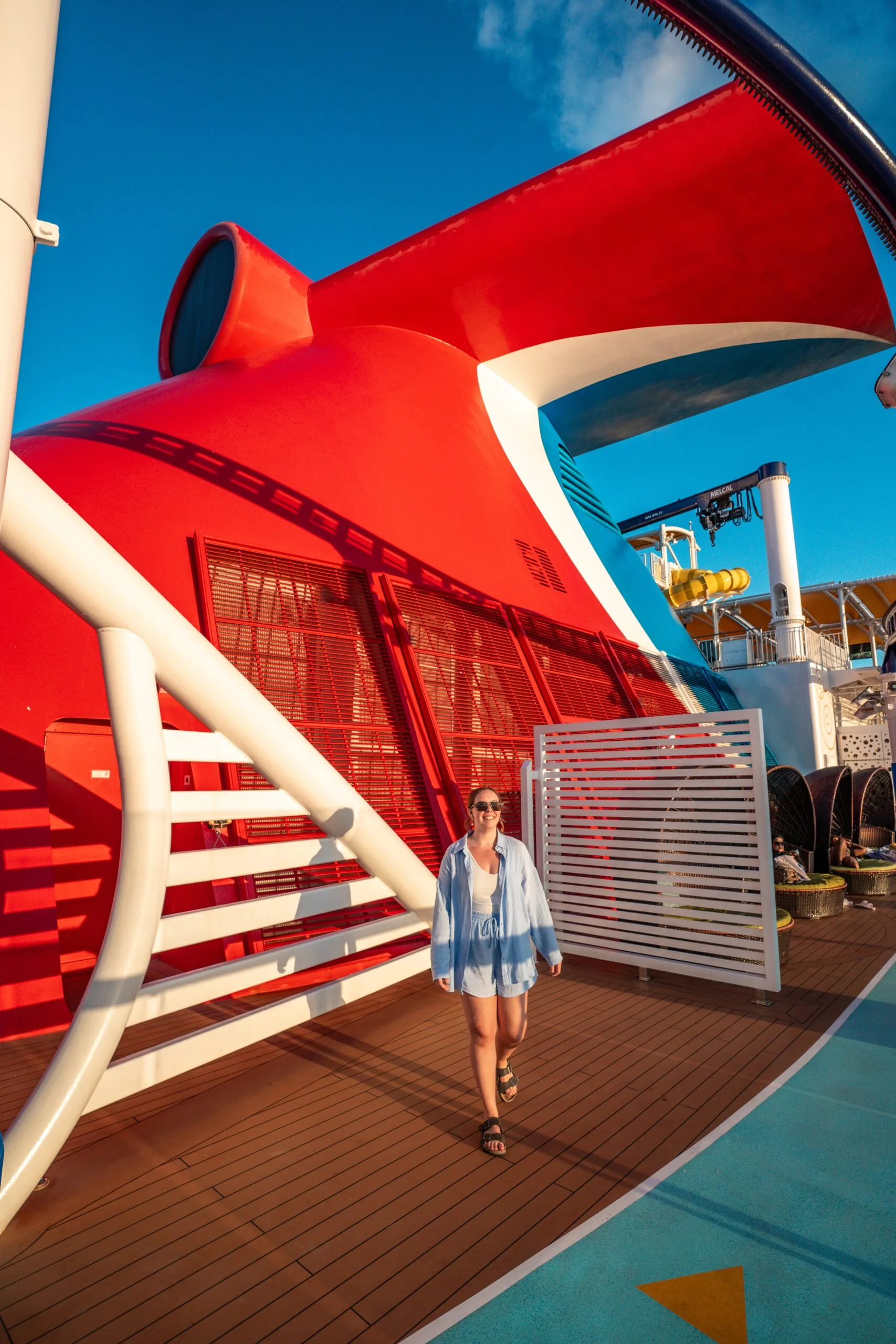 A person in sunglasses and blue attire walks on a cruise ship deck under a clear sky, near a large red smokestack, conveying leisure and vacation vibes.