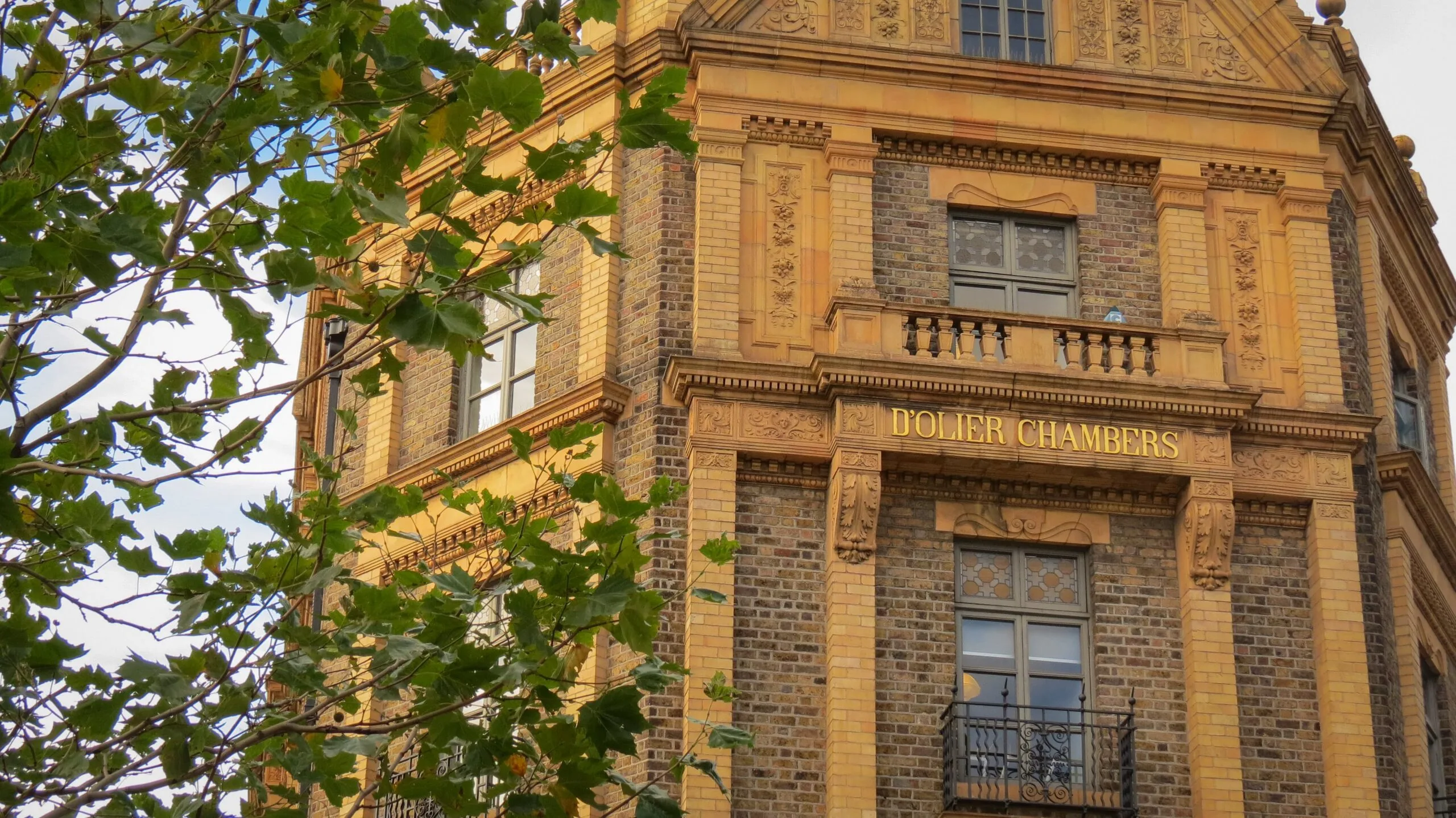 Brick building facade with ornate stonework and gold lettering reading "D'Olier Chambers." Green leaves partially frame the scene against a blue sky in dublin for a weekend.