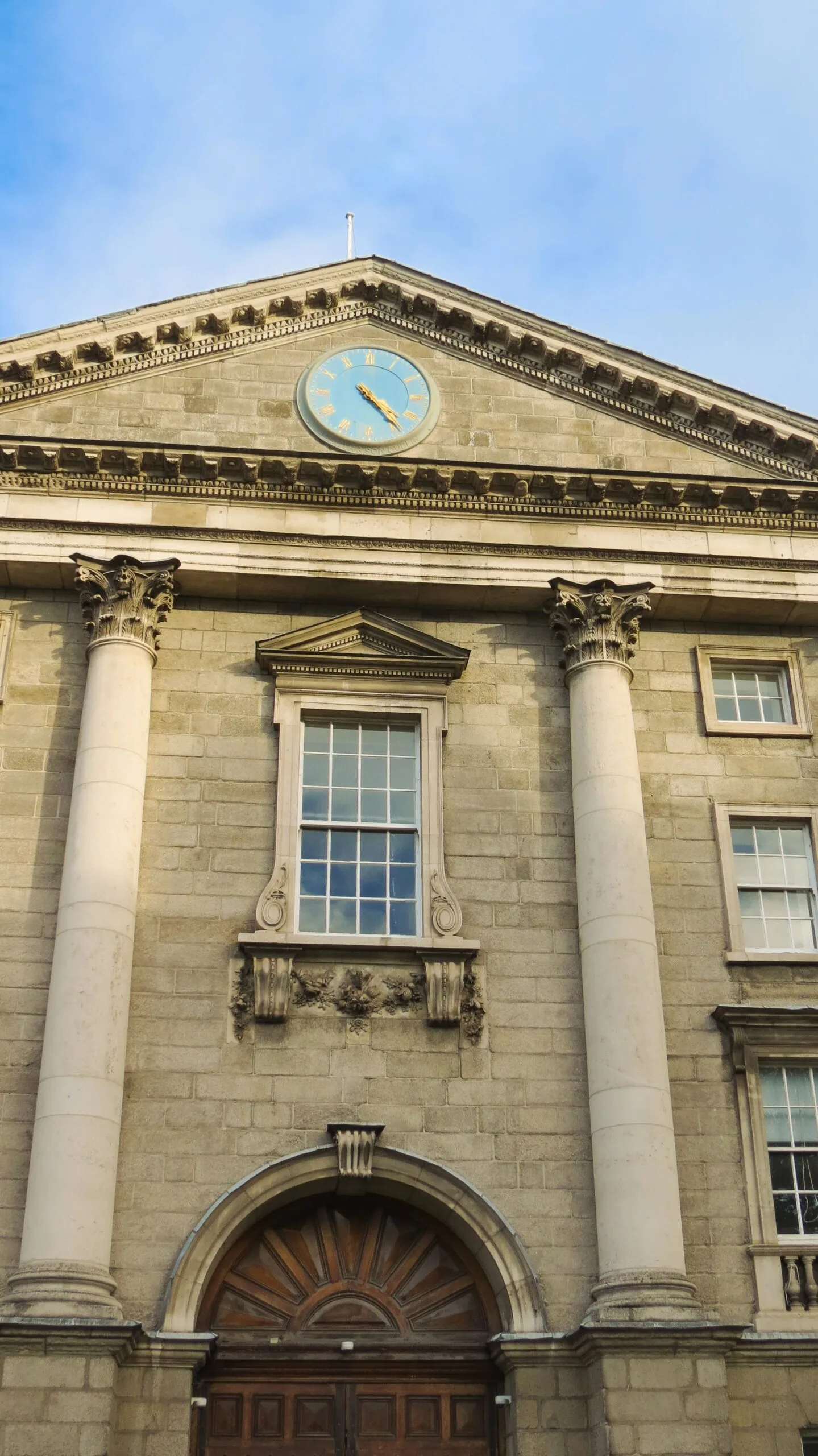 Historic stone building facade with tall columns and arched wooden door. A blue clock with gold Roman numerals sits atop the structure. Bright, clear sky.
