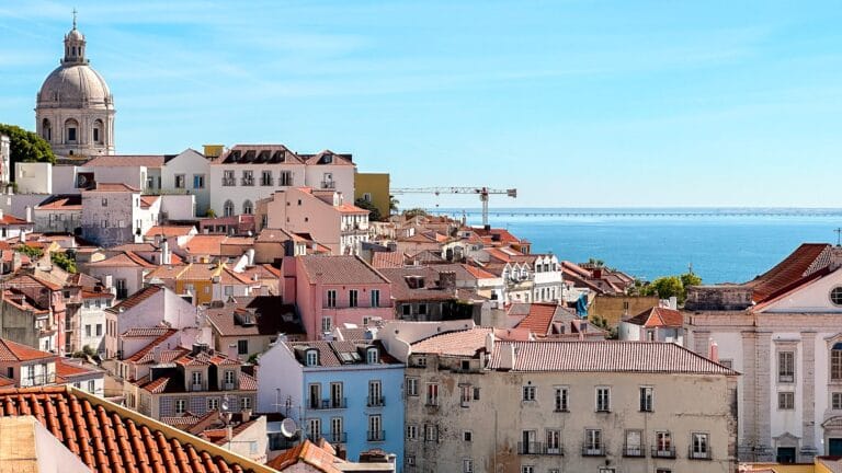 Scenic view of Lisbon's rooftops featuring colorful buildings and a large dome. The ocean is visible in the background under a clear blue sky.