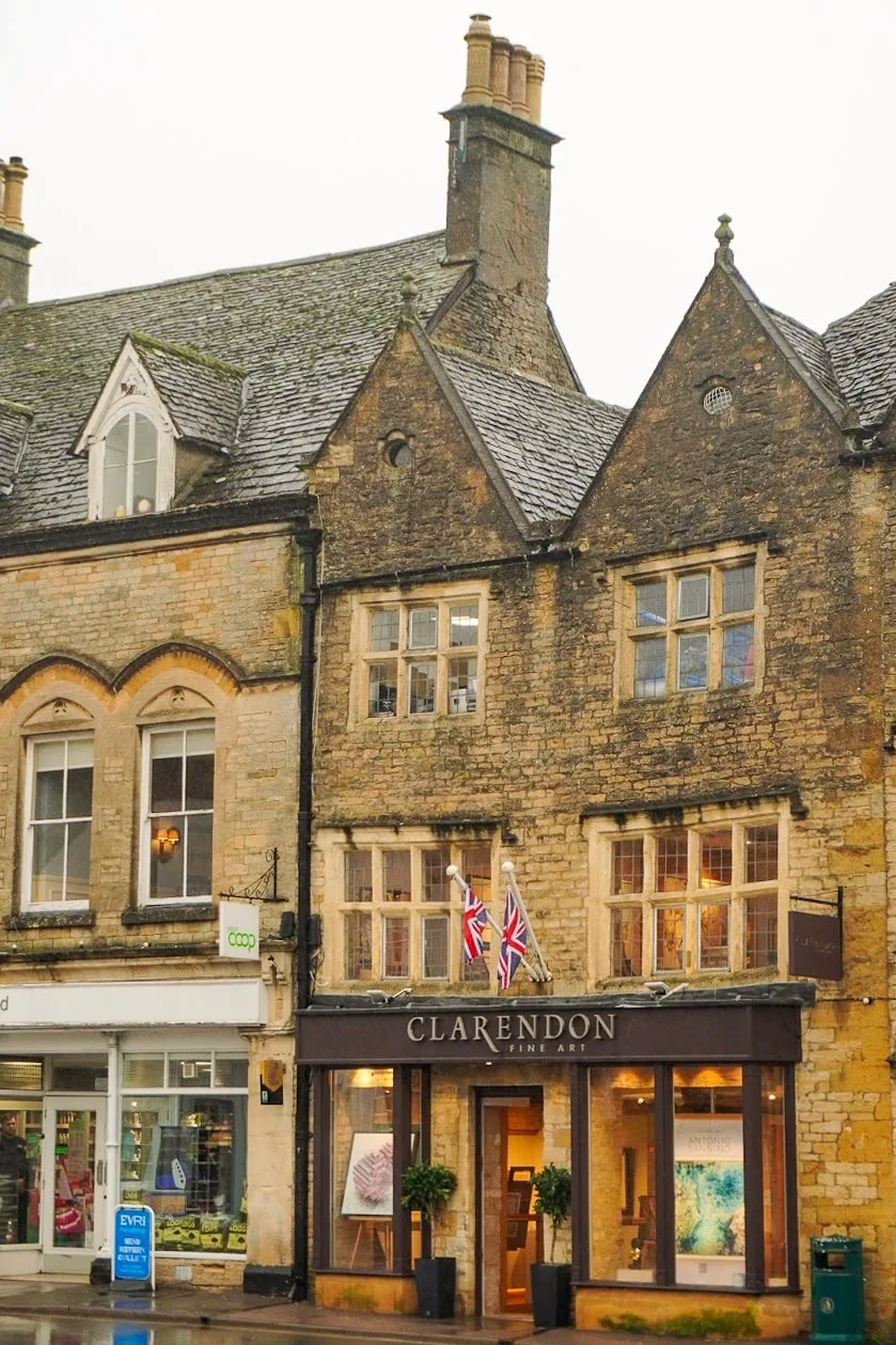Historic stone building in Stow on the Wold UK with steep gabled roofs, housing "Clarendon Fine Art." Two Union Jack flags are displayed; a neighboring co-op store is visible.