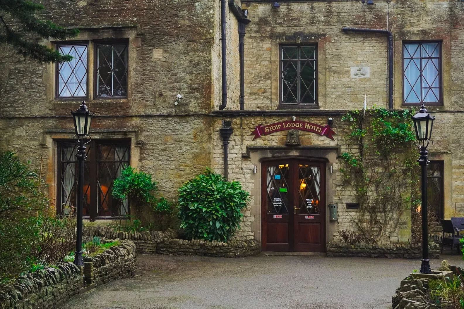 Ancient stone building with ivy, featuring diamond-paned windows and a wooden door labeled "Stow Lodge Hotel." Tall lanterns flank the entrance, creating a cozy feel.