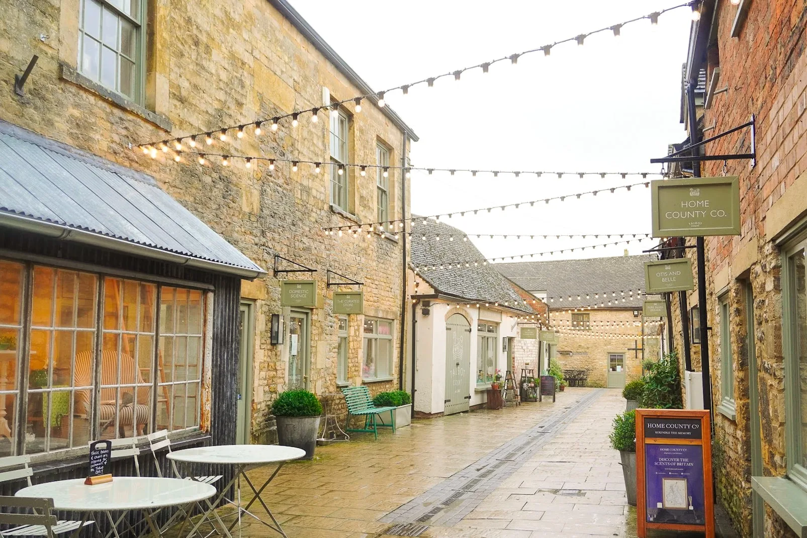 Charming cobblestone alley with rustic stone buildings adorned with string lights. Outdoor tables and shop signs create a quaint, inviting atmosphere.