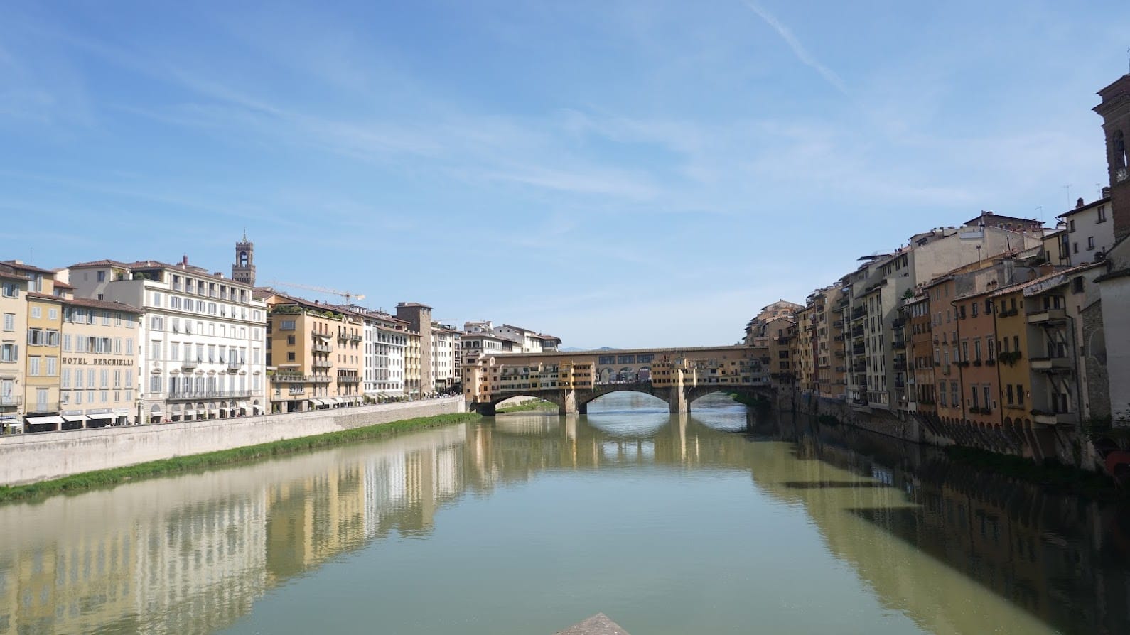 A serene view of Ponte Vecchio in Florence, Italy, with historic buildings lining the Arno River under a clear blue sky. The calm water reflects the structures.