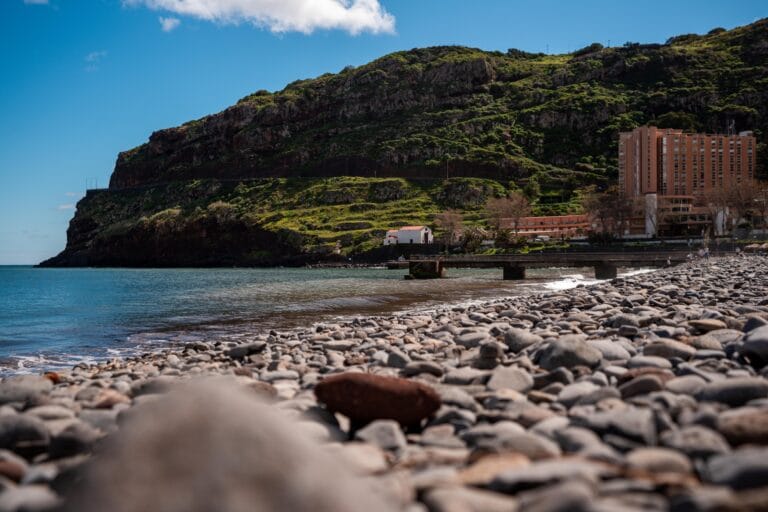 Rocky beach scene with calm blue ocean, green mountain cliffs, and a distant hotel under a clear blue sky. Peaceful and inviting coastal view.