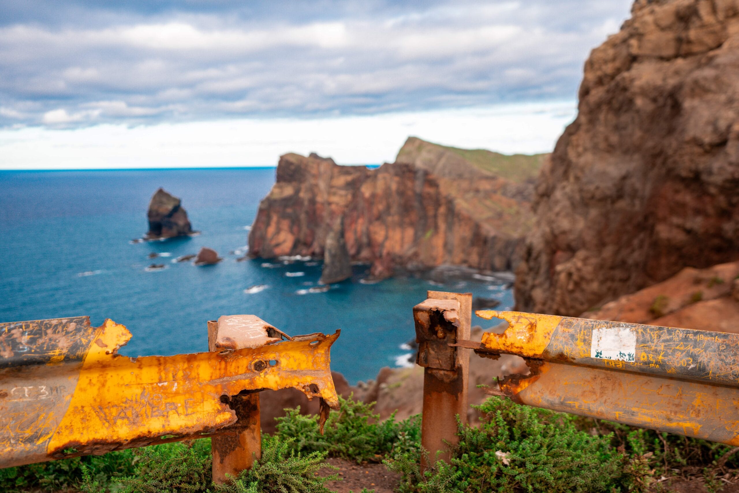 Rusty metal guardrail contrasts with vibrant cliffs and ocean in the background. Overcast sky and rugged landscape create a dramatic mood.