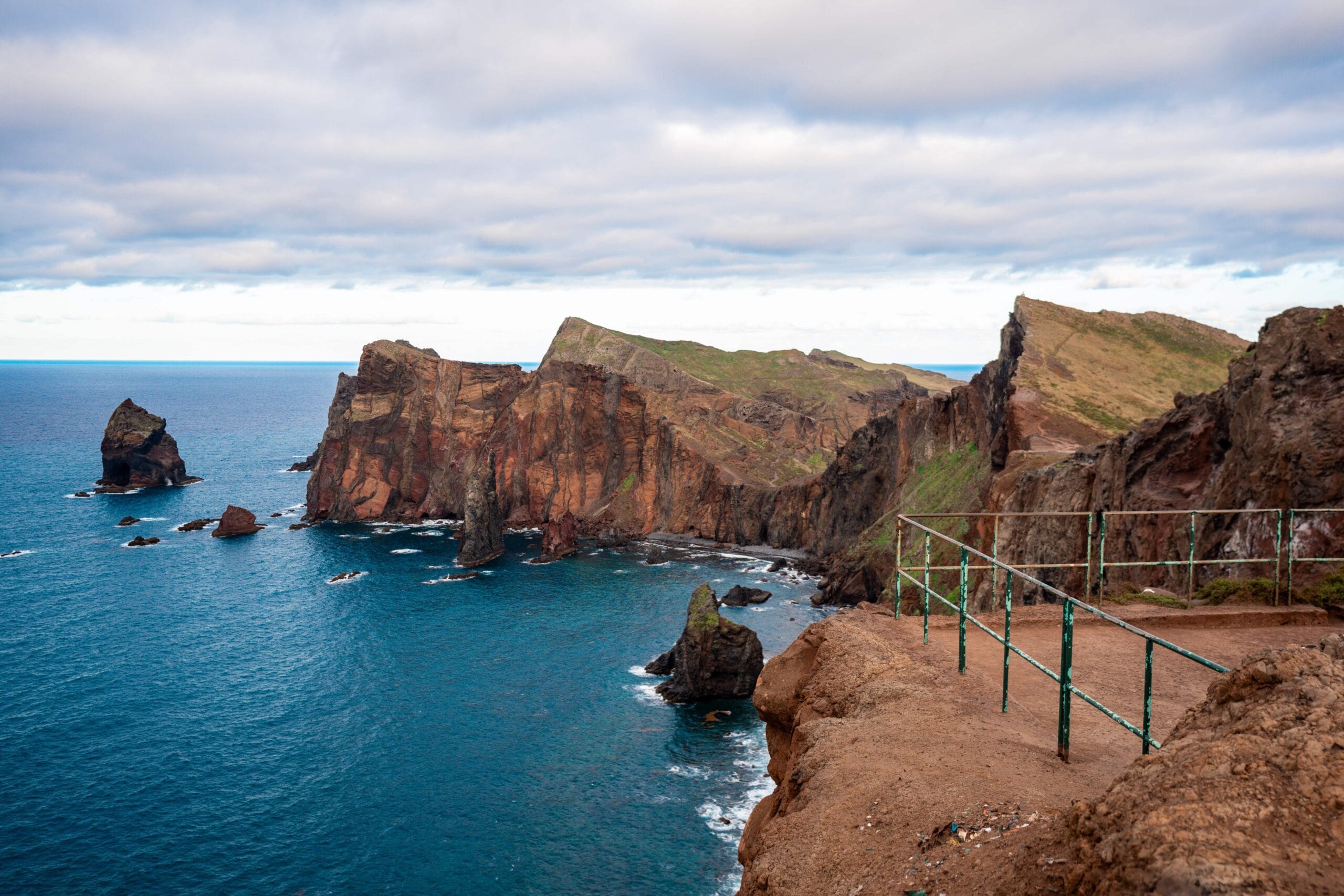 Cliffside view rugged, reddish-brown rock formations in Madeira Portugal jutting into a blue ocean under a cloudy sky, with a metal railing in the foreground