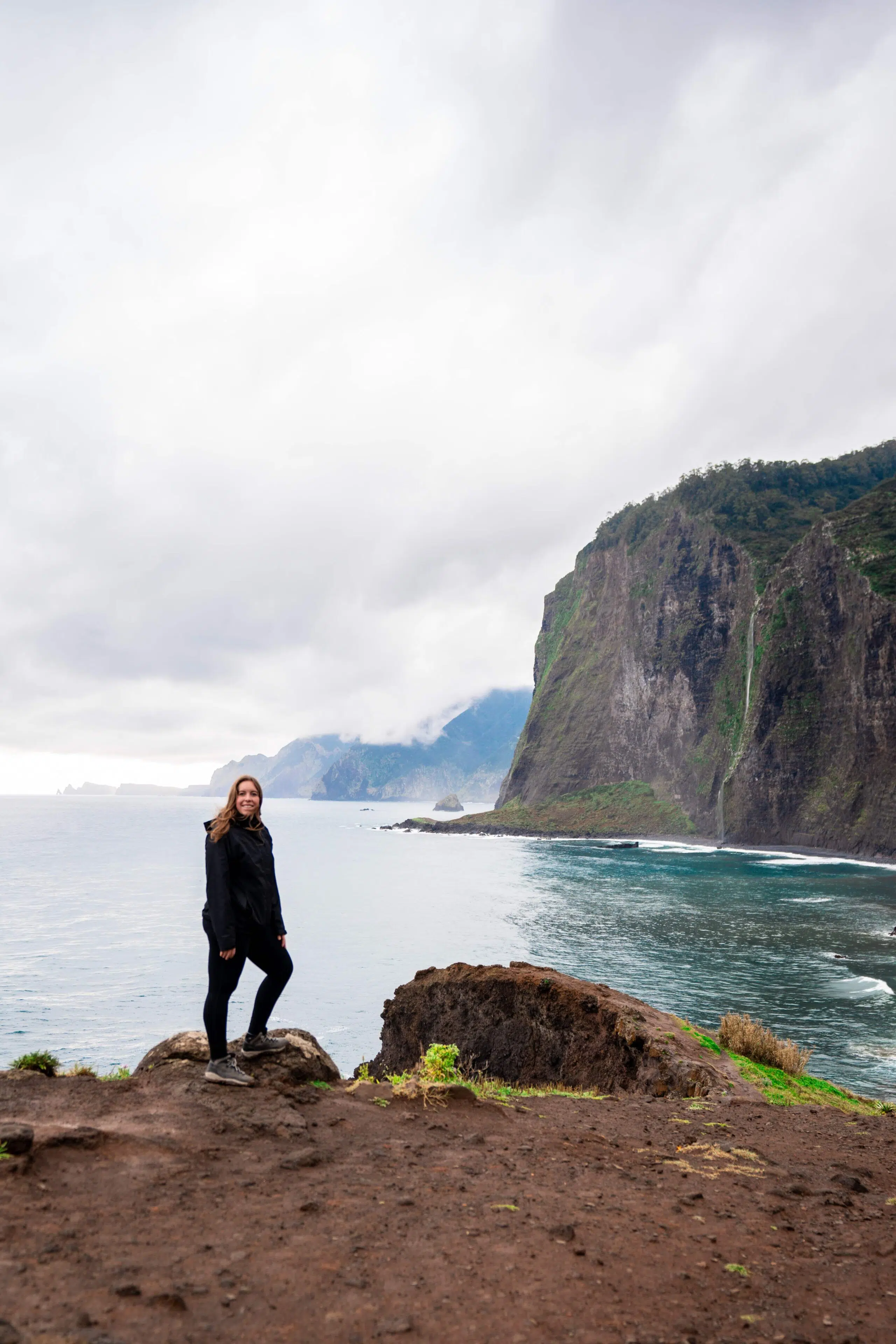 A woman in a black jacket smiles while standing on a rocky cliff overlooking a vast ocean. Cloudy skies and rugged cliffs create a serene, adventurous mood.