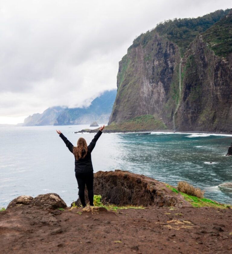 15 Amazing Things to See and Do in Madeira Portugal 15 A woman stands on a cliff, arms raised in triumph, overlooking the ocean. Towering cliffs and a cloudy sky create a dramatic and empowering scene.