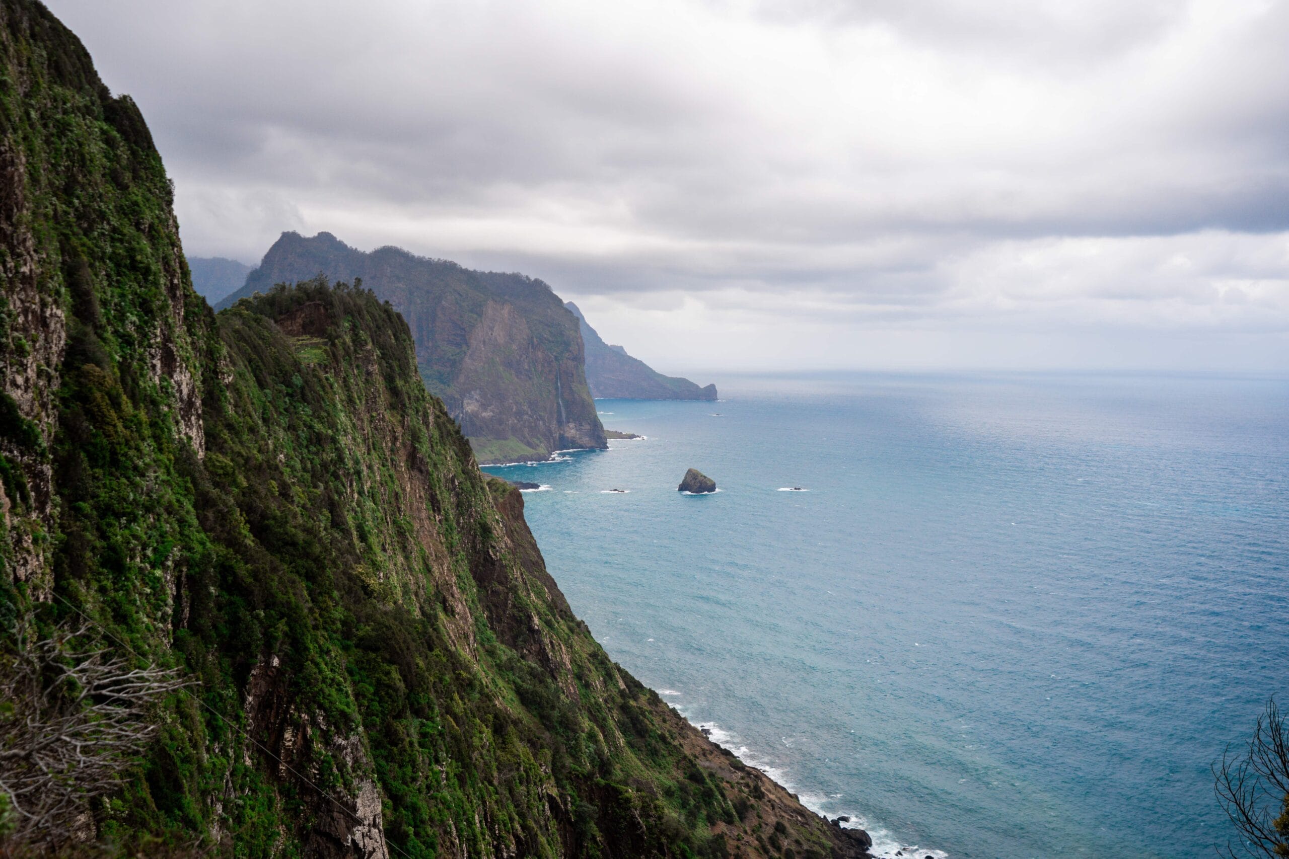 Steep green cliffs descend to a calm, blue ocean under a cloudy sky. The rugged coastline and distant mountains evoke a serene, remote atmosphere.