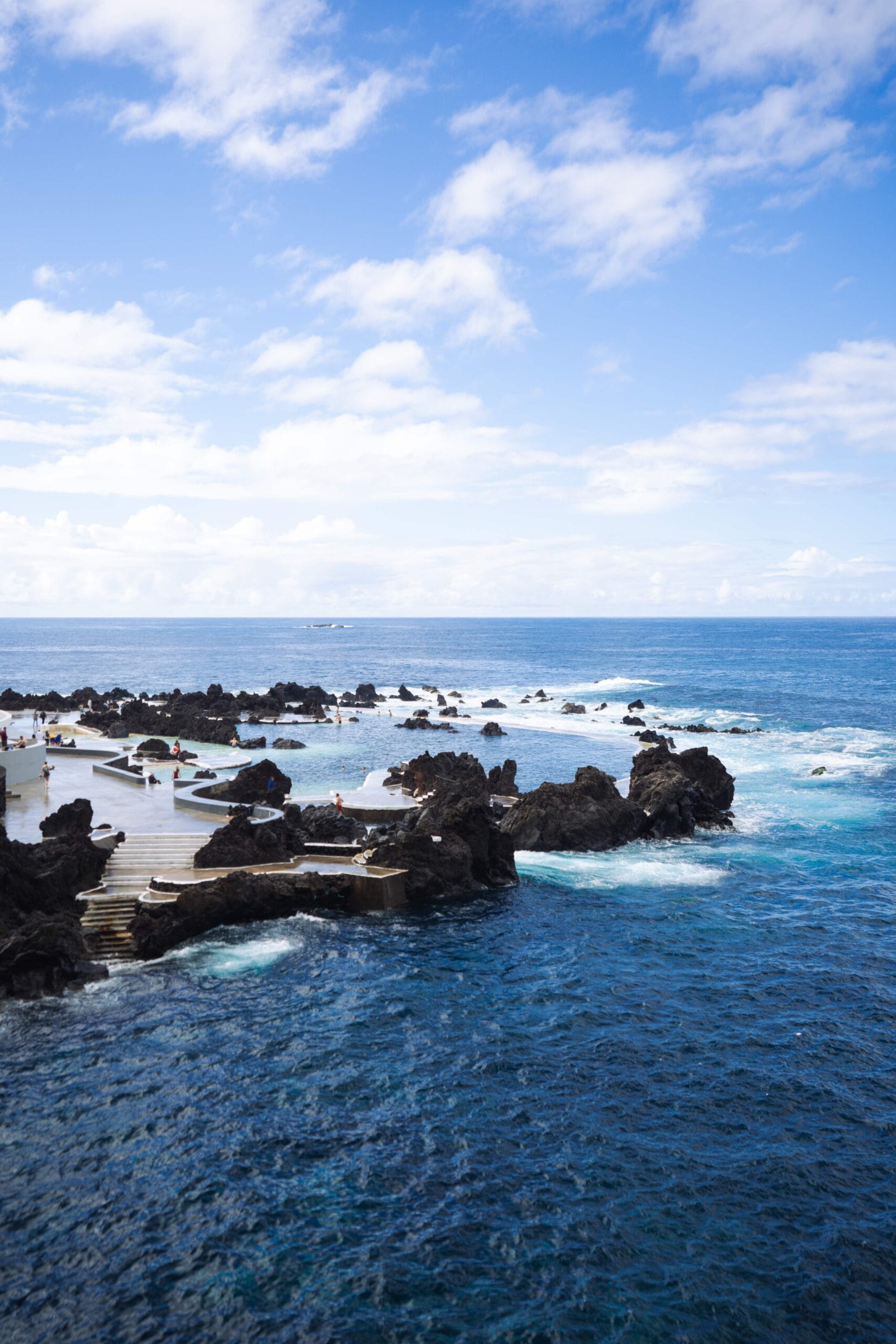 Coastal scene with dark volcanic rocks, turquoise ocean waves, and a blue sky with scattered clouds. People are enjoying a natural swimming area.